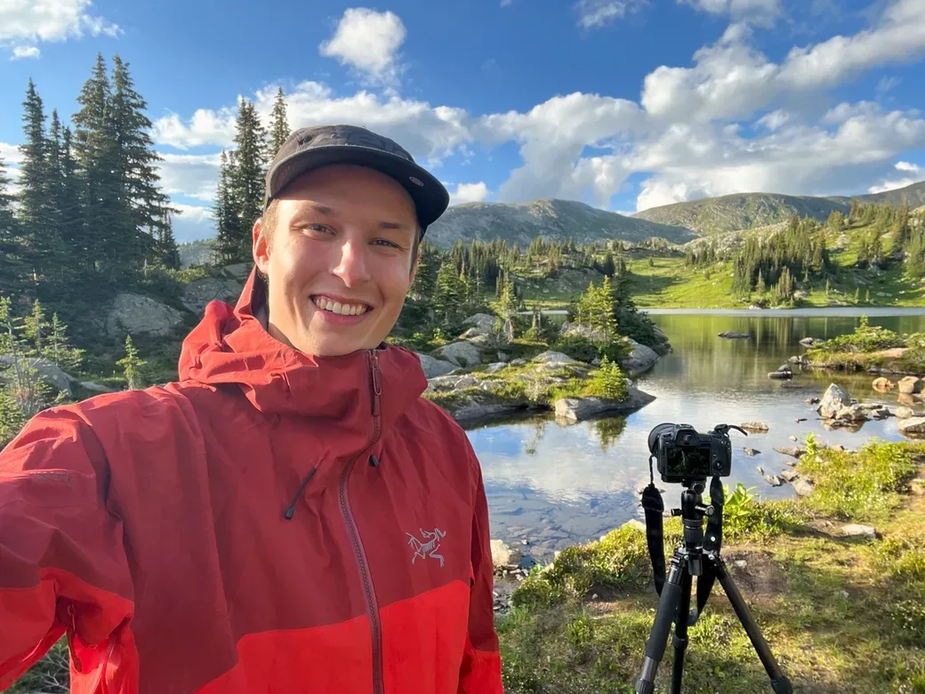 Brendan smiling in front of a mountain lake with his camera on a tripod